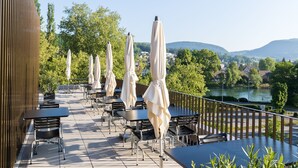 Een terras met zitplaatsen buiten, parasols en uitzicht op een rivier en heuvels.