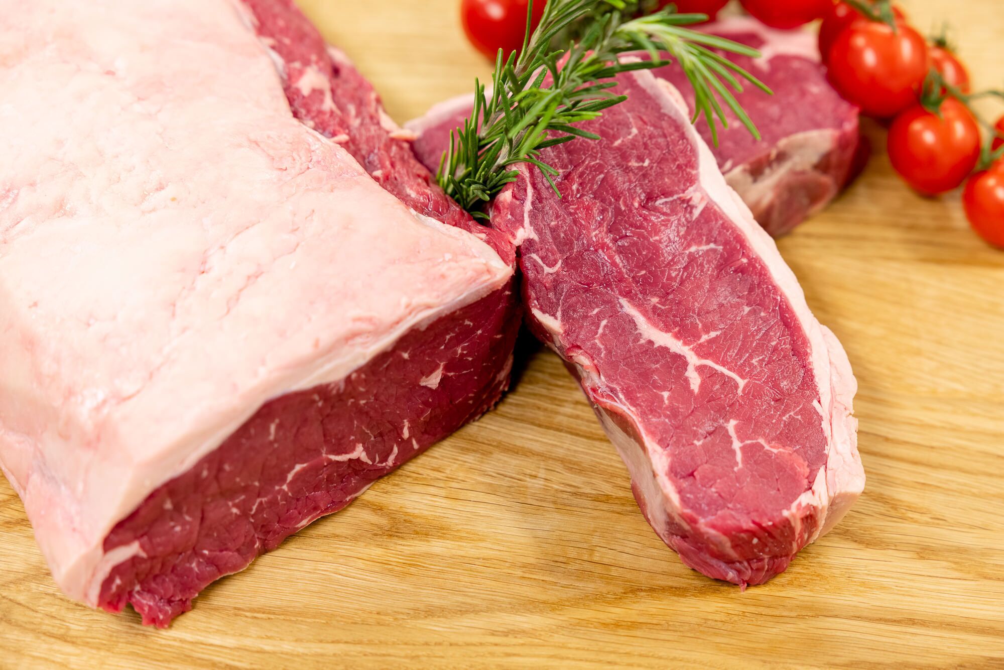 Deux steaks crus sur une planche à découper en bois, avec une branche de romarin et des tomates cerises en arrière-plan.