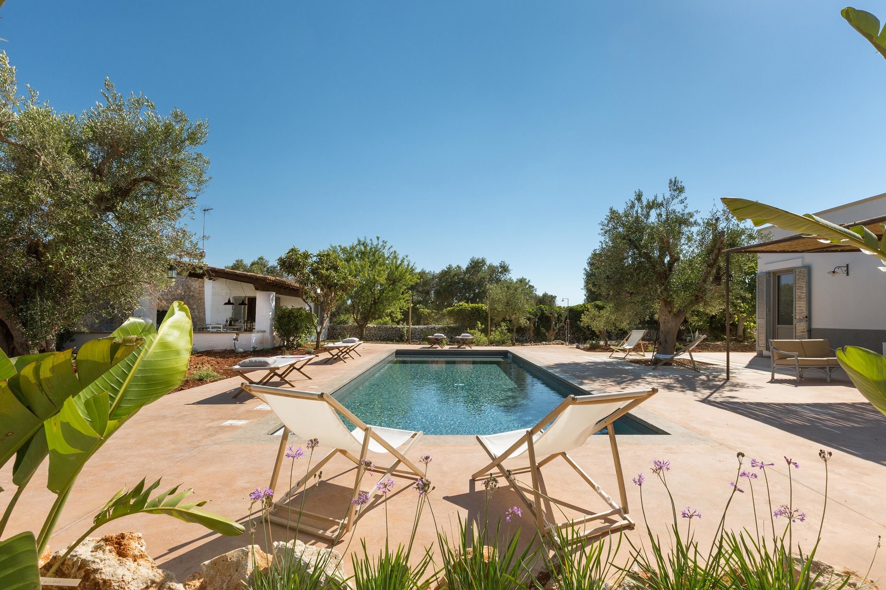A rectangular swimming pool with lounge chairs and a small building in the background.