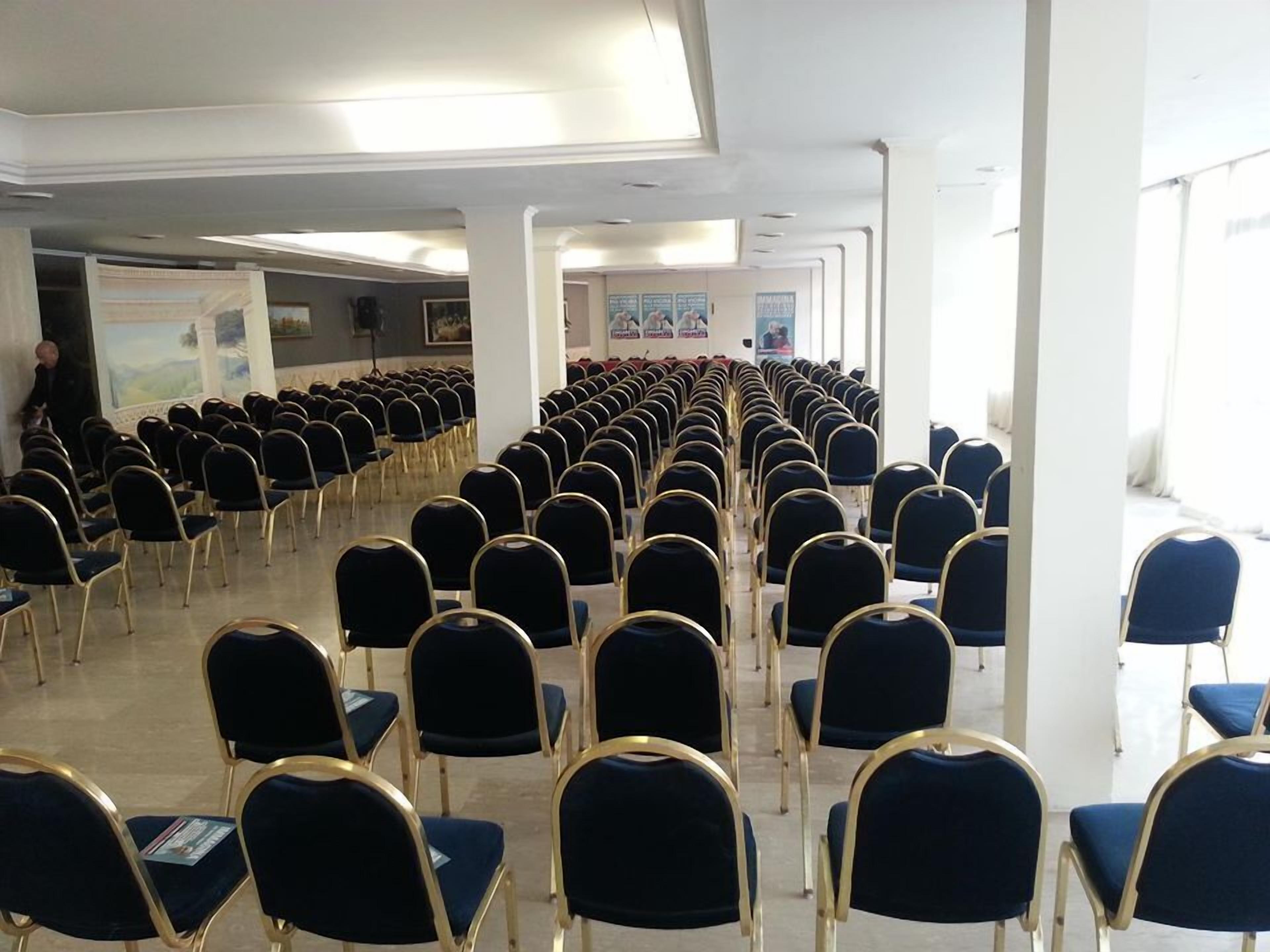 A conference room with rows of blue chairs and gold frames, white pillars, and a large window on the right.
