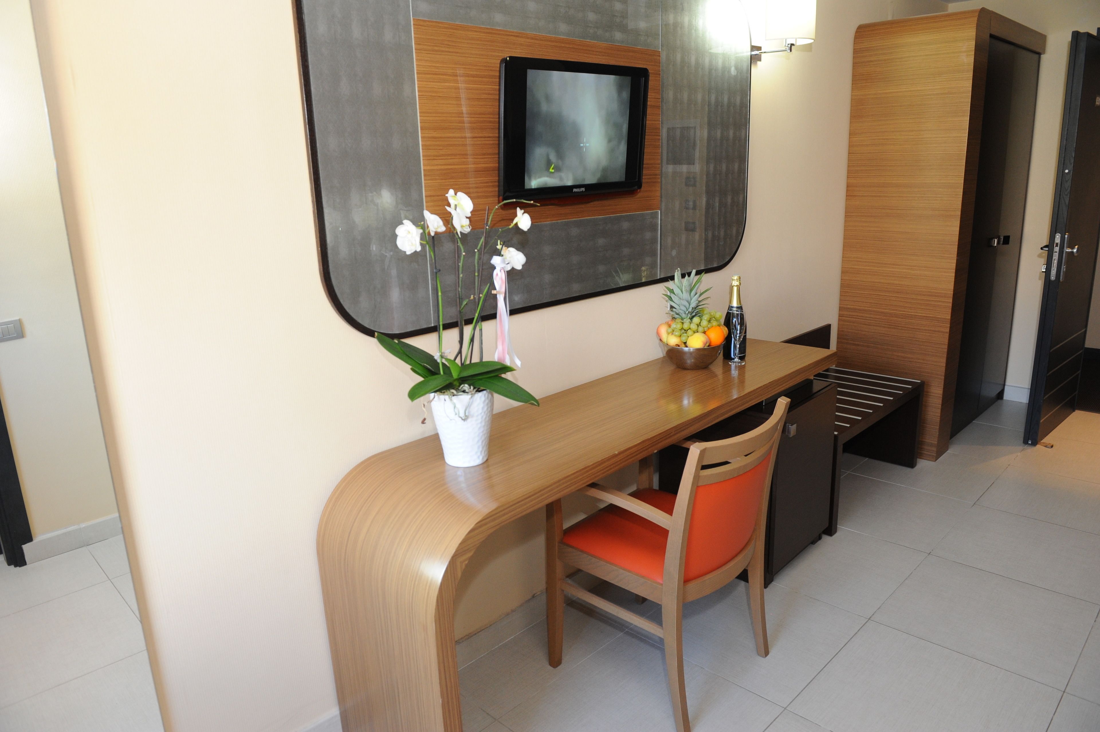 A compact hotel room with a wooden desk, a chair, a small potted plant, a fruit bowl, and a television mounted on the wall.