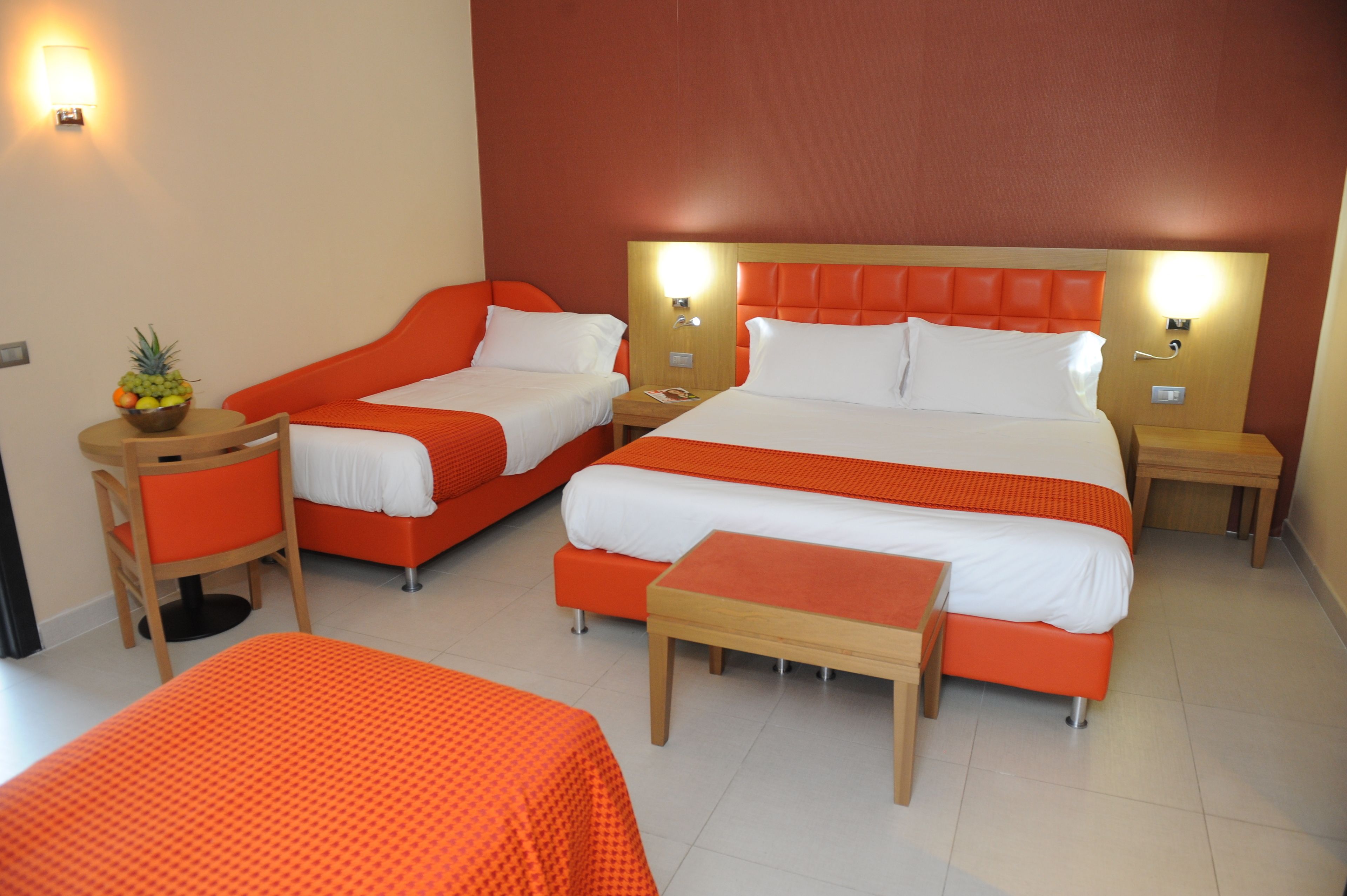 Une chambre d’hôtel avec deux lits simples, un lit double et un canapé-lit. On y trouve une petite table avec un panier de fruits, une chaise et une table de chevet avec une lampe.