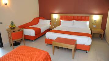 Une chambre d’hôtel avec deux lits simples, un lit double et un canapé-lit. On y trouve une petite table avec un panier de fruits, une chaise et une table de chevet avec une lampe.