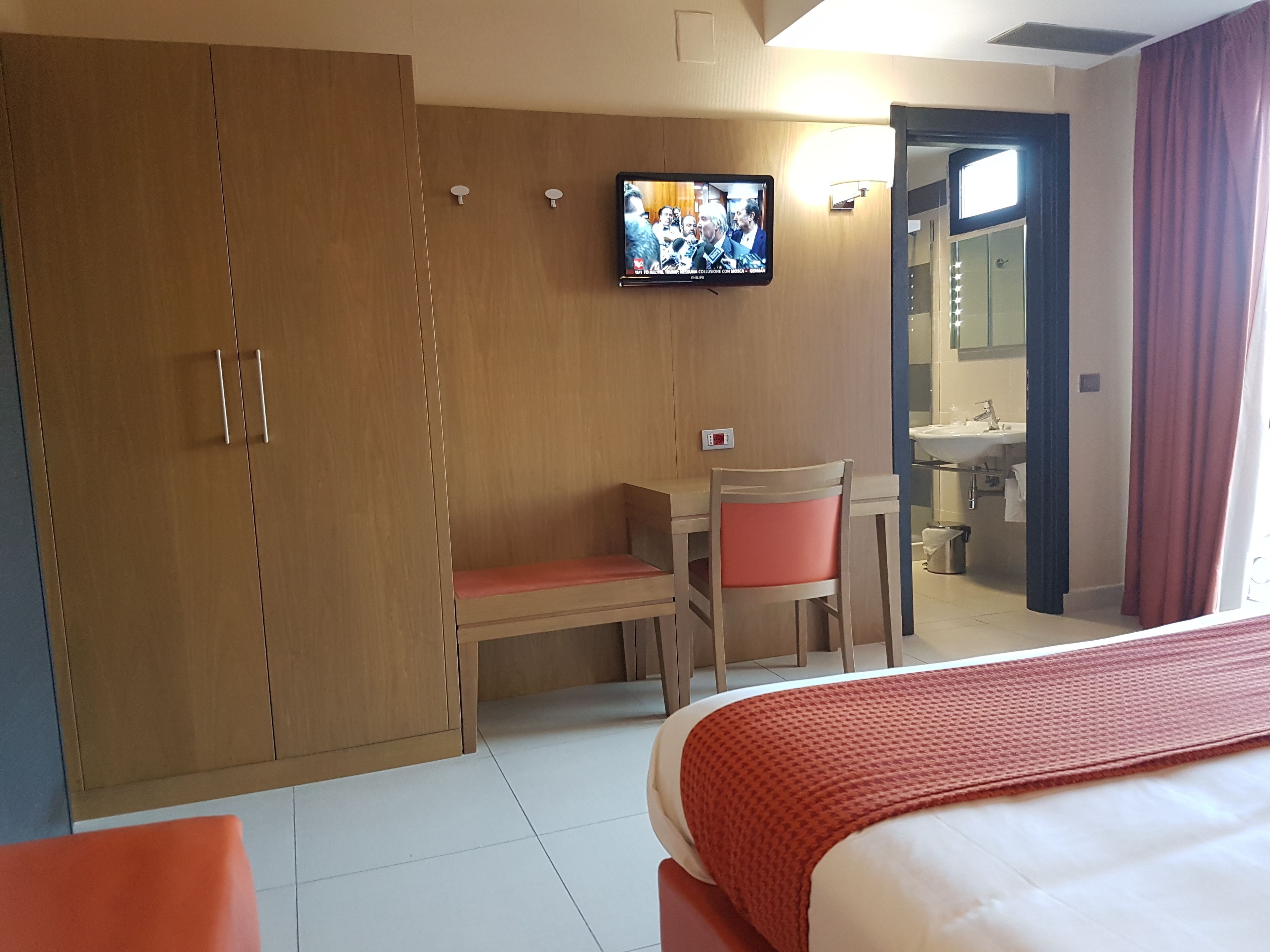 A hotel room with a bed, a desk with a chair, a television mounted on the wall, and a bathroom visible through an open door.