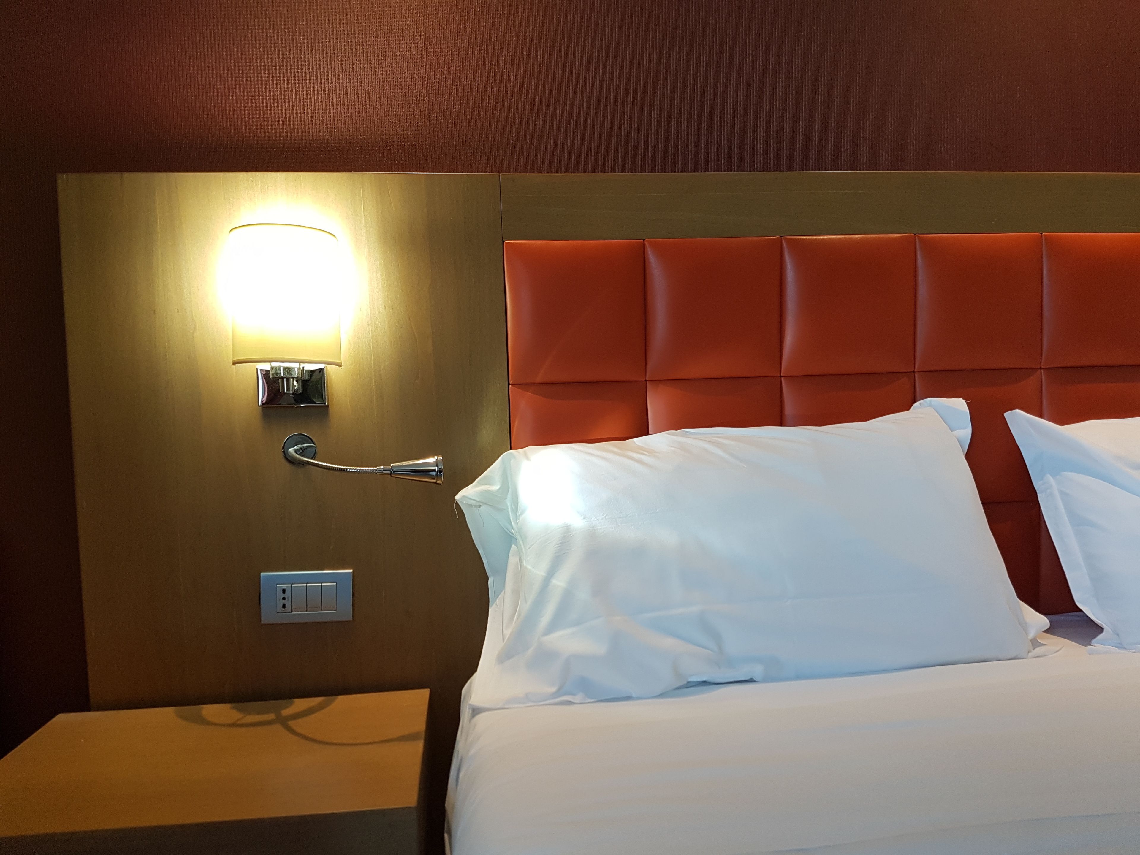 Una habitación de hotel con cama, almohadas blancas, mesita de noche, luz de pared y termostato.
