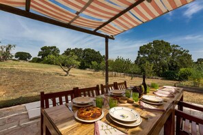 Une table à manger installée à l’extérieur, avec des assiettes, des verres et de la nourriture, sous une marquise à rayures, offrant une vue sur des arbres et un champ d’herbe.