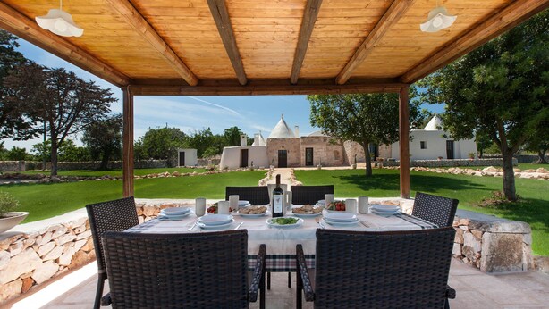 Un área de comedor al aire libre con una pérgola de madera, una mesa preparada para cuatro personas y una vista de un edificio histórico al fondo.