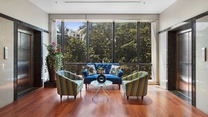 A modern lobby with a blue sofa, two patterned armchairs, a glass coffee table, and a large window offering a view of trees and buildings. - Hotel Satkar Grande (Thane)