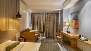 Una habitación de hotel con cama, escritorio, silla, una mesita y un cuadro en la pared.