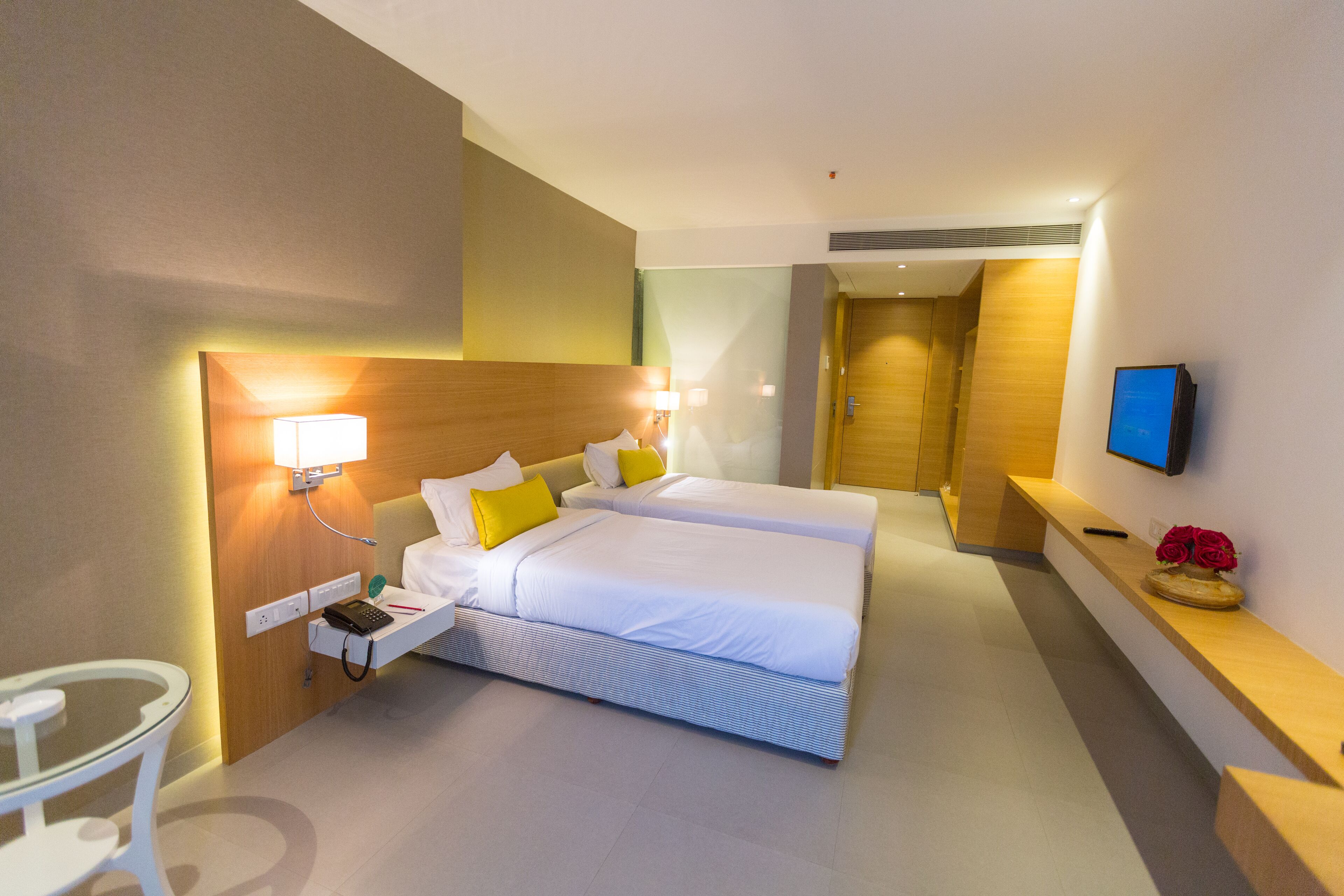 Habitación de hotel con dos camas, una mesita de noche con lámpara, un televisor y una pequeña mesa con un jarrón de flores.