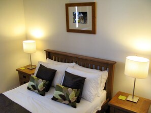 Un lit bien fait, avec du linge de lit blanc et deux coussins décoratifs, flanqué de deux tables de chevet sur lesquelles reposent des lampes et des blocs-notes.