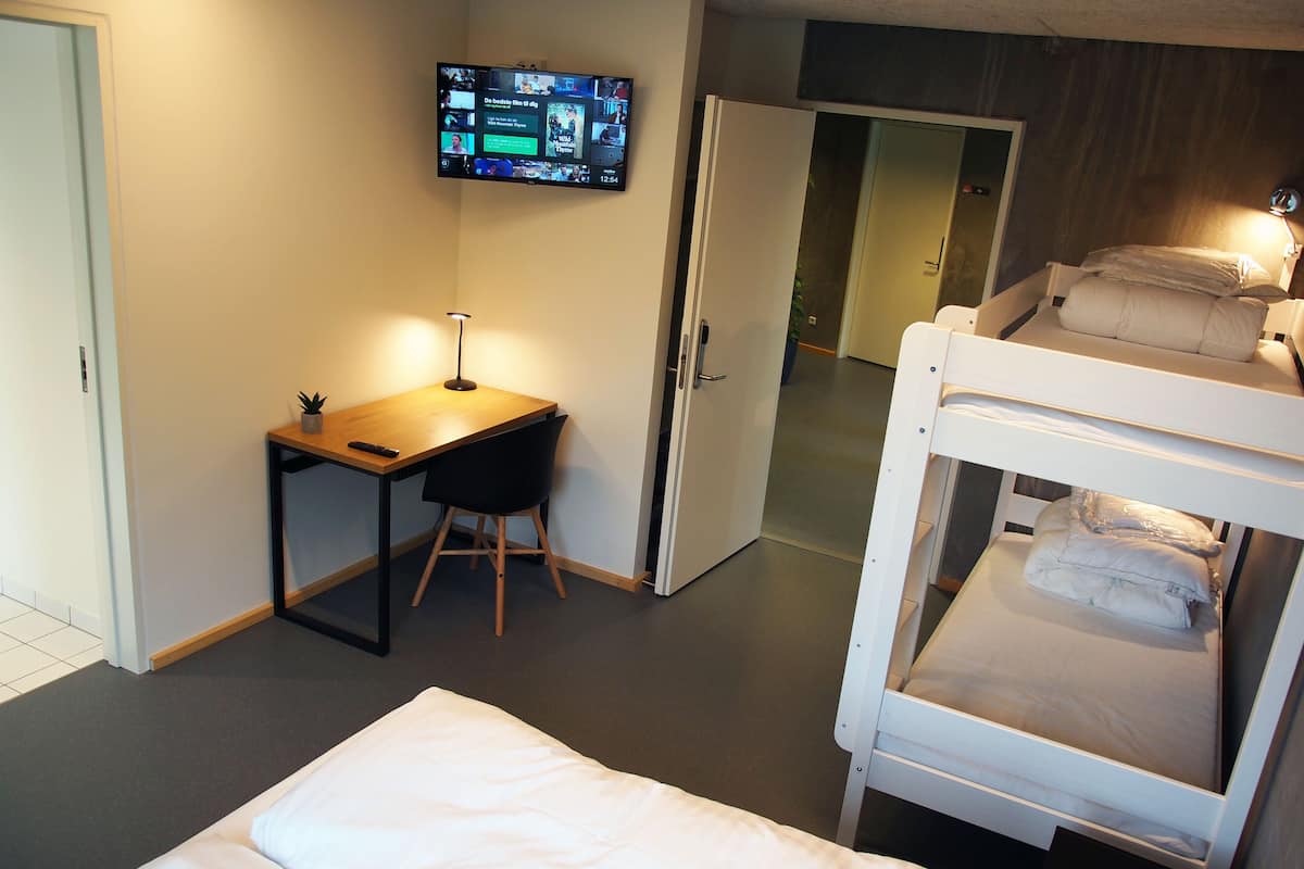Habitación de hotel con una cama, un escritorio con silla, un televisor montado en la pared y una puerta que da a otra habitación.