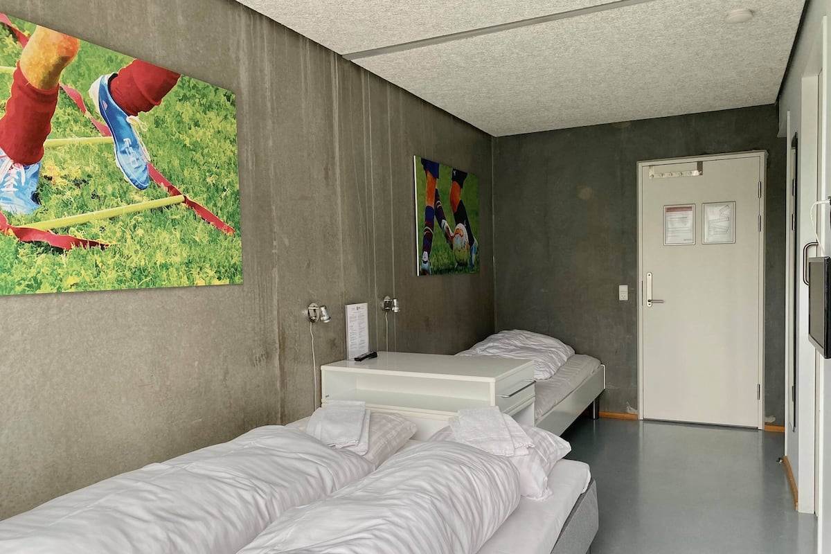 Habitación de hotel con dos camas individuales, una mesita de noche pequeña, un espejo y una puerta con un cartel.