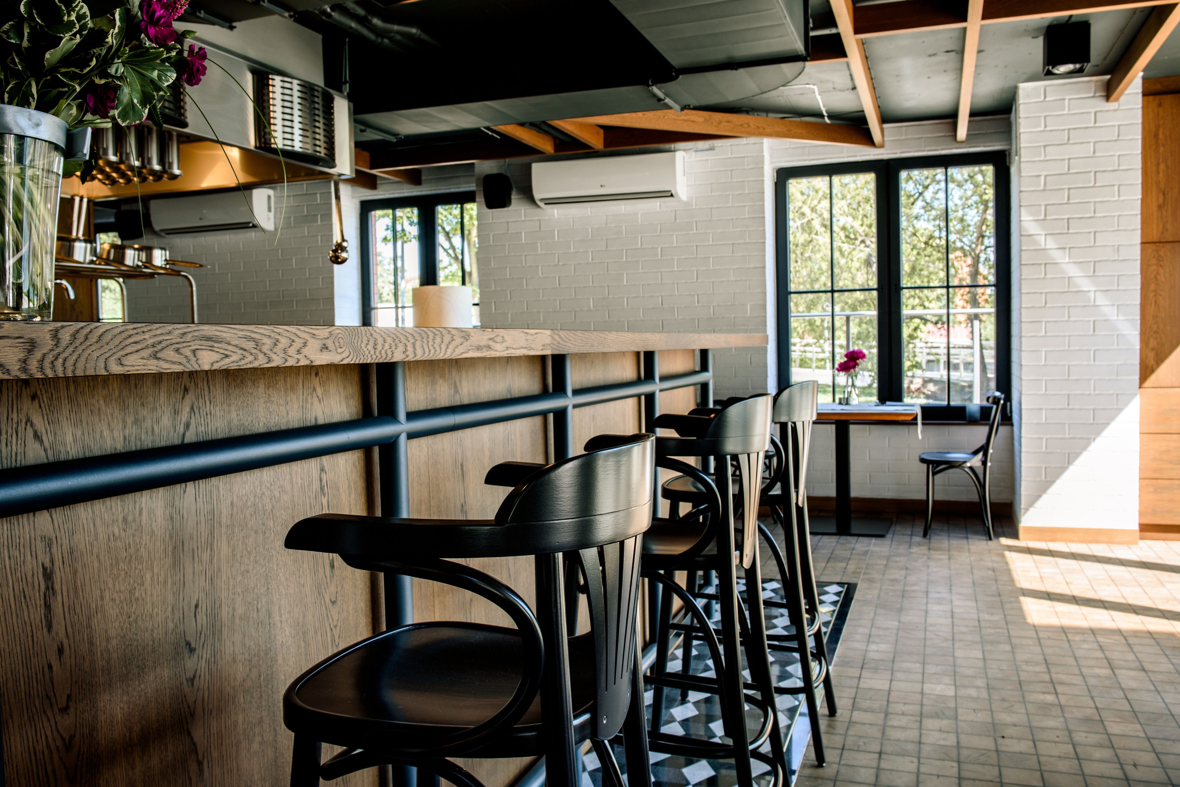 A bar area with wooden counter and black bar stools, a window with a view of trees, and a brick wall.
