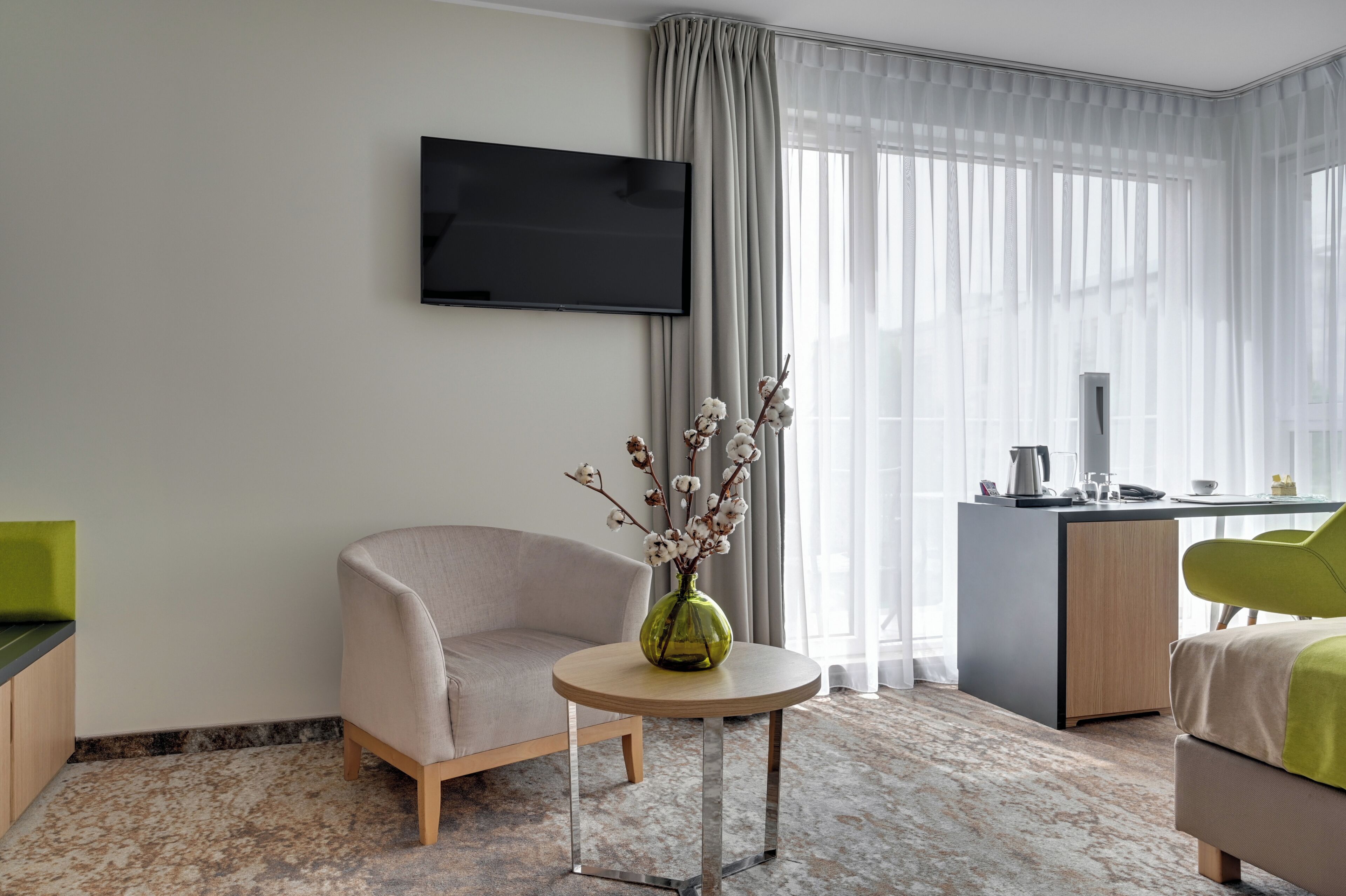 A hotel room with a sofa, a small round table with a vase, a desk with a chair, a TV mounted on the wall, and a large window with sheer curtains.