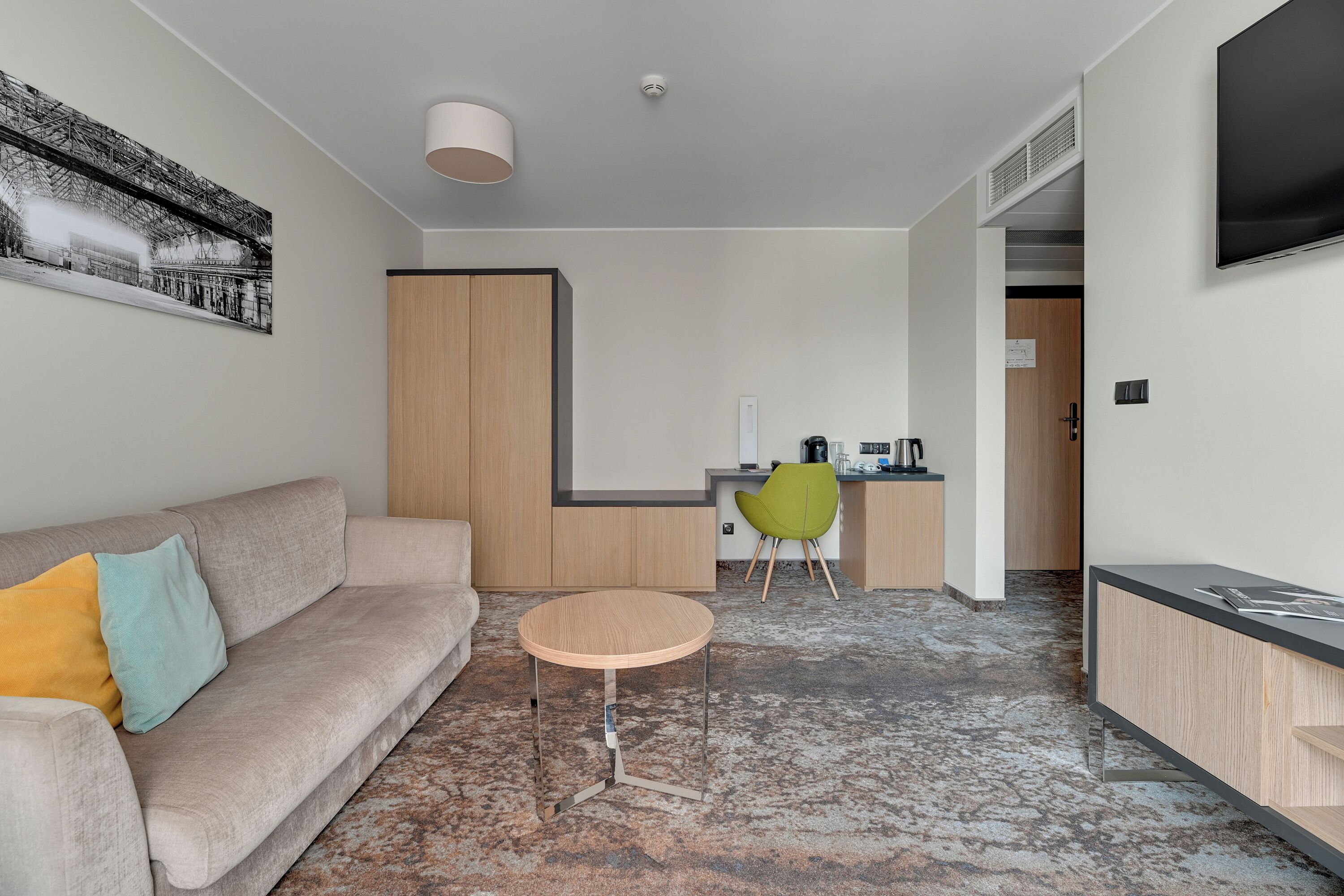 Una habitación de hotel moderna con sofá, una mesa redonda, una silla verde, una zona de cocina y un televisor montado en la pared.