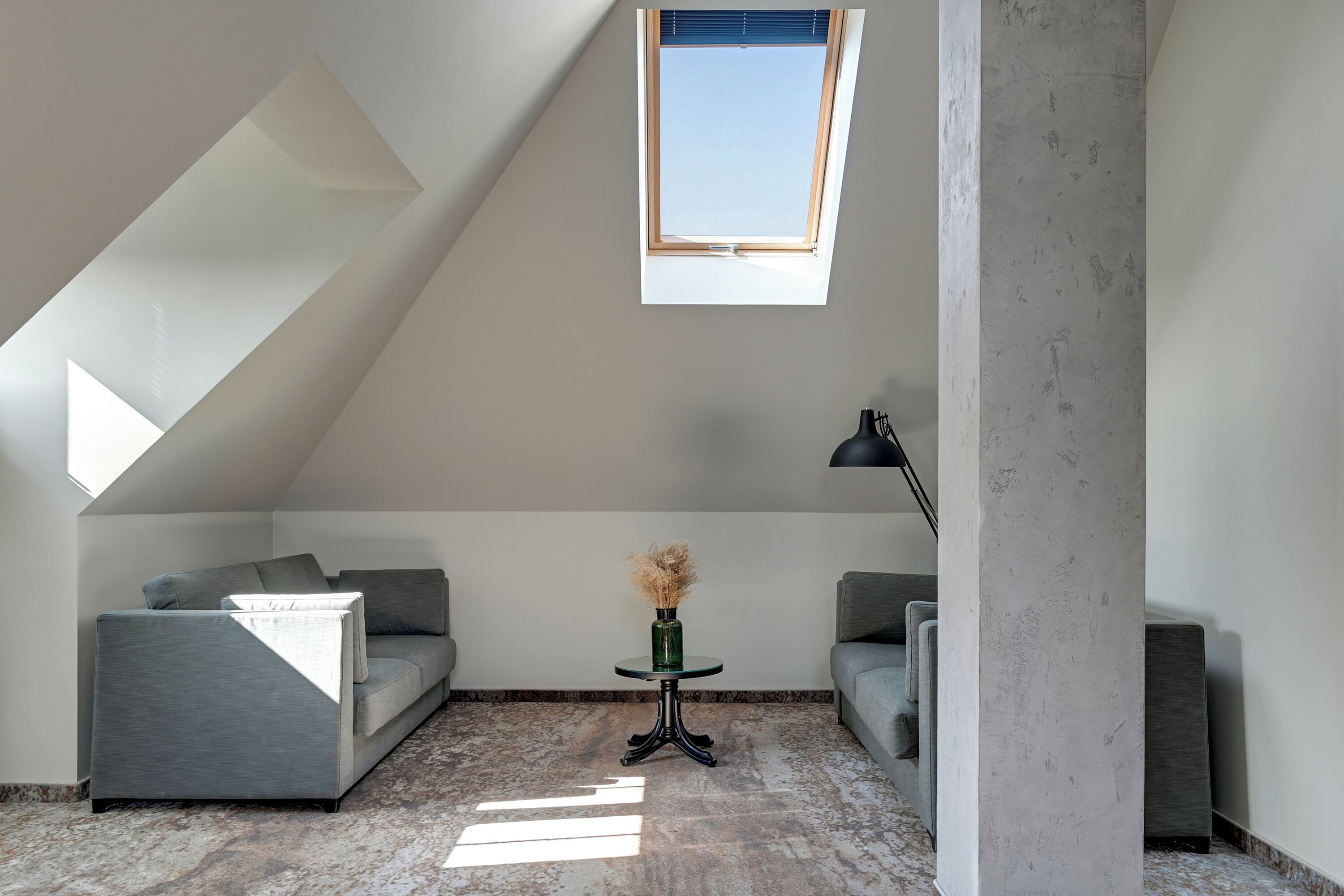 A modern loft bedroom with a sloped ceiling, a skylight, a gray sofa, a small round table with a vase, and a lamp.