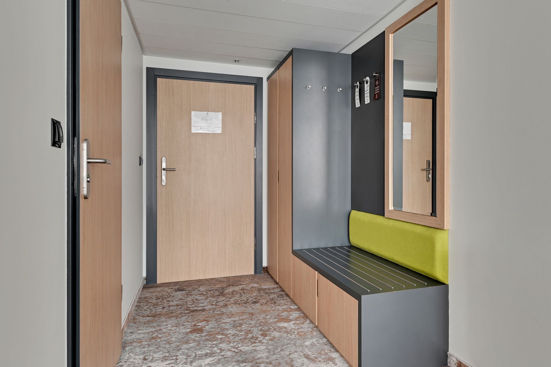 A hallway with a wooden door, a grey bench with a green cushion, and a mirror on the wall.