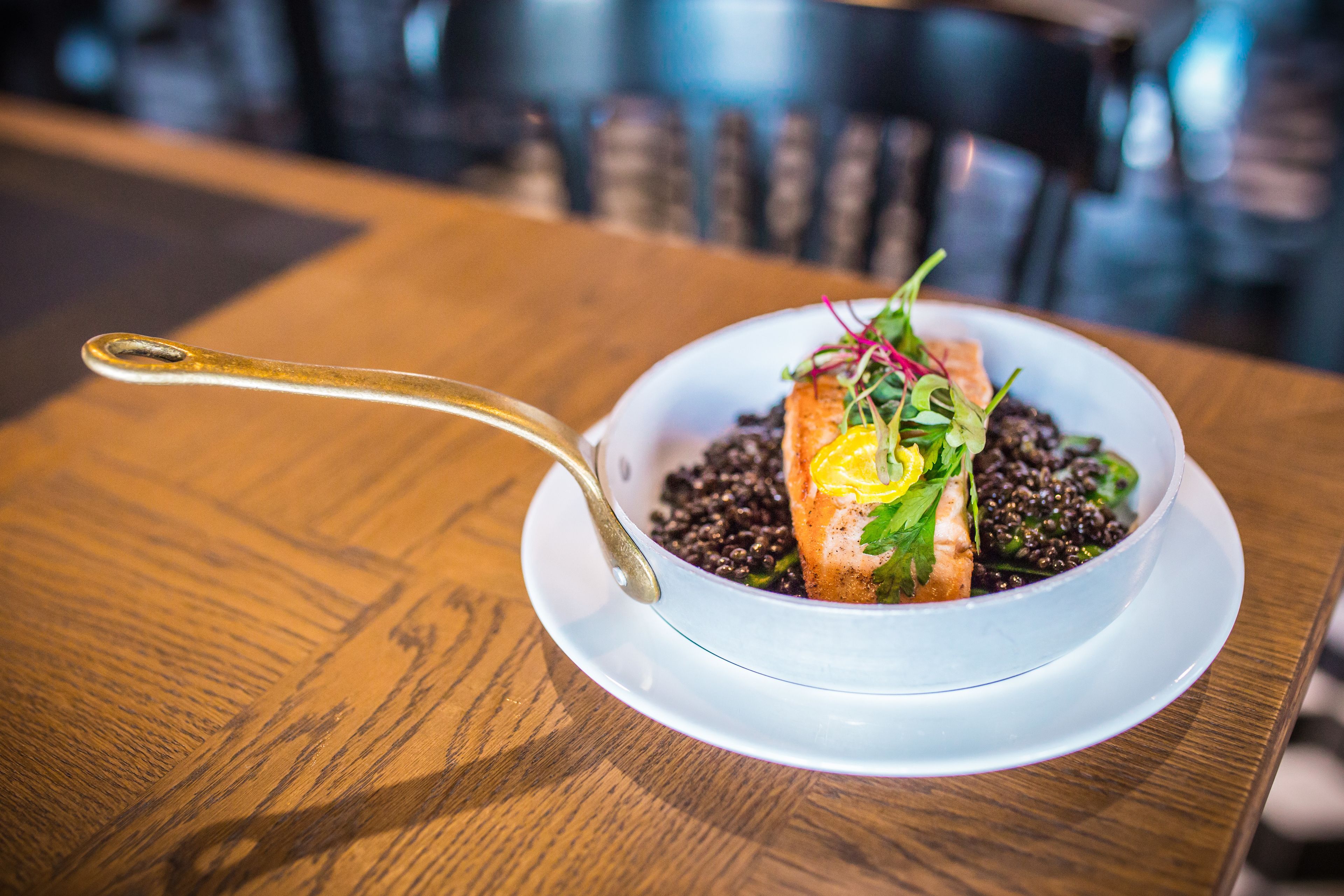A white bowl with a salmon fillet, black beans, and greens on a wooden table.