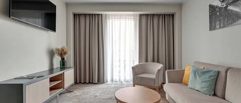 A modern living room with a flat-screen TV, a sofa, a chair, a small round table, and a large window with curtains.
