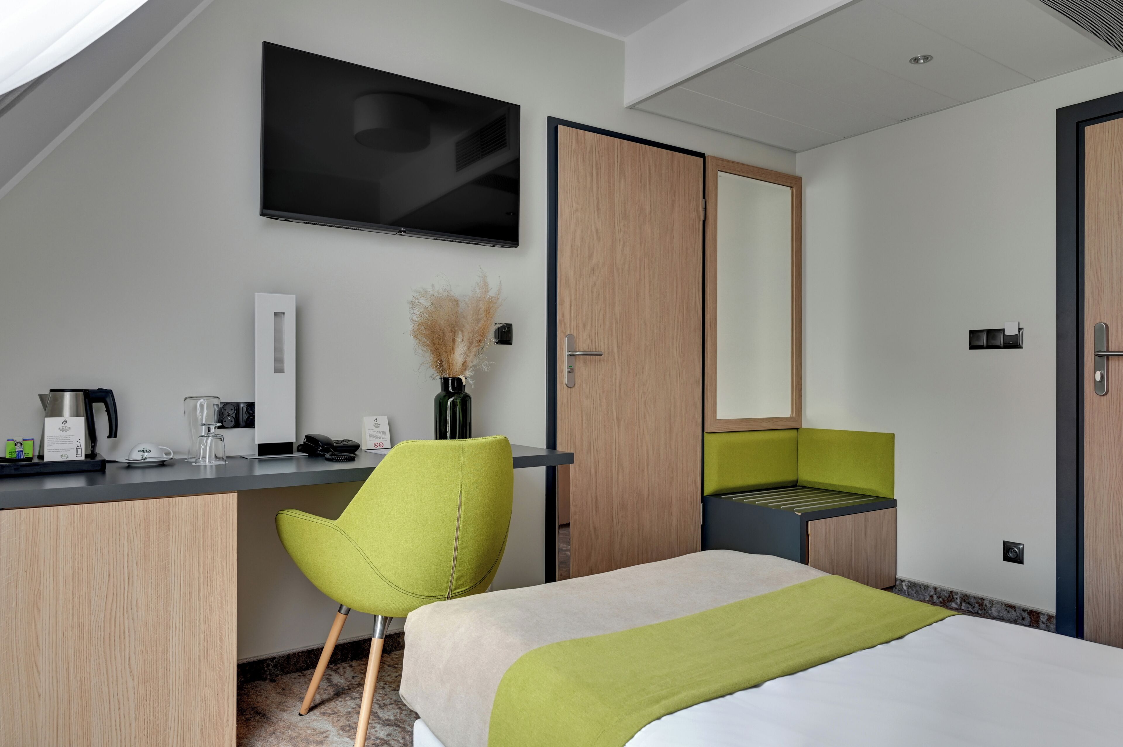 A modern hotel room with a bed, desk, chair, TV, and a bathroom with a mirror and sink.