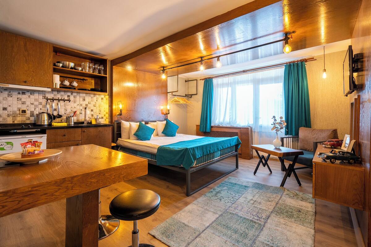 A compact hotel room with a kitchenette, a bed with teal bedding, a small dining table, and a seating area.