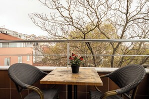 Un balcon avec une table et deux chaises, un pot de fleurs avec des fleurs rouges, et une vue sur des immeubles et des arbres.