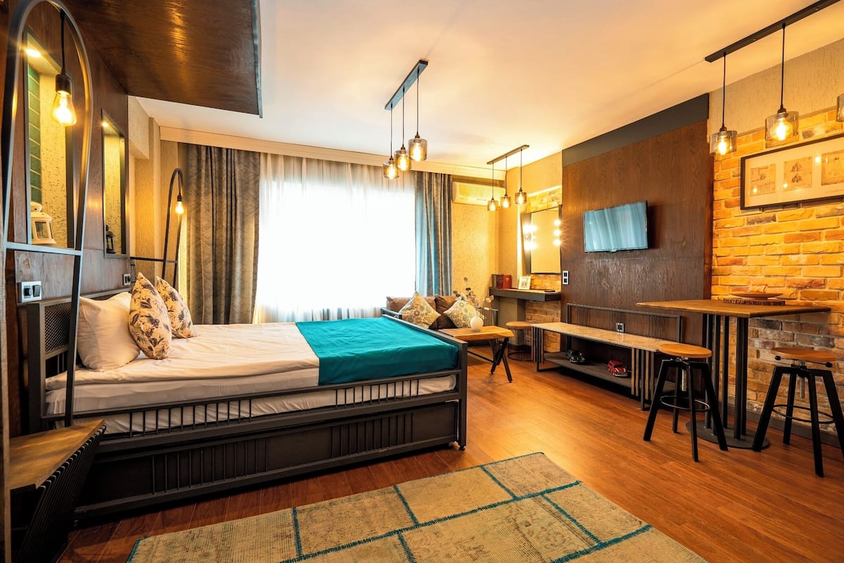 A modern hotel room with a bed, a desk, a TV, and a brick wall.