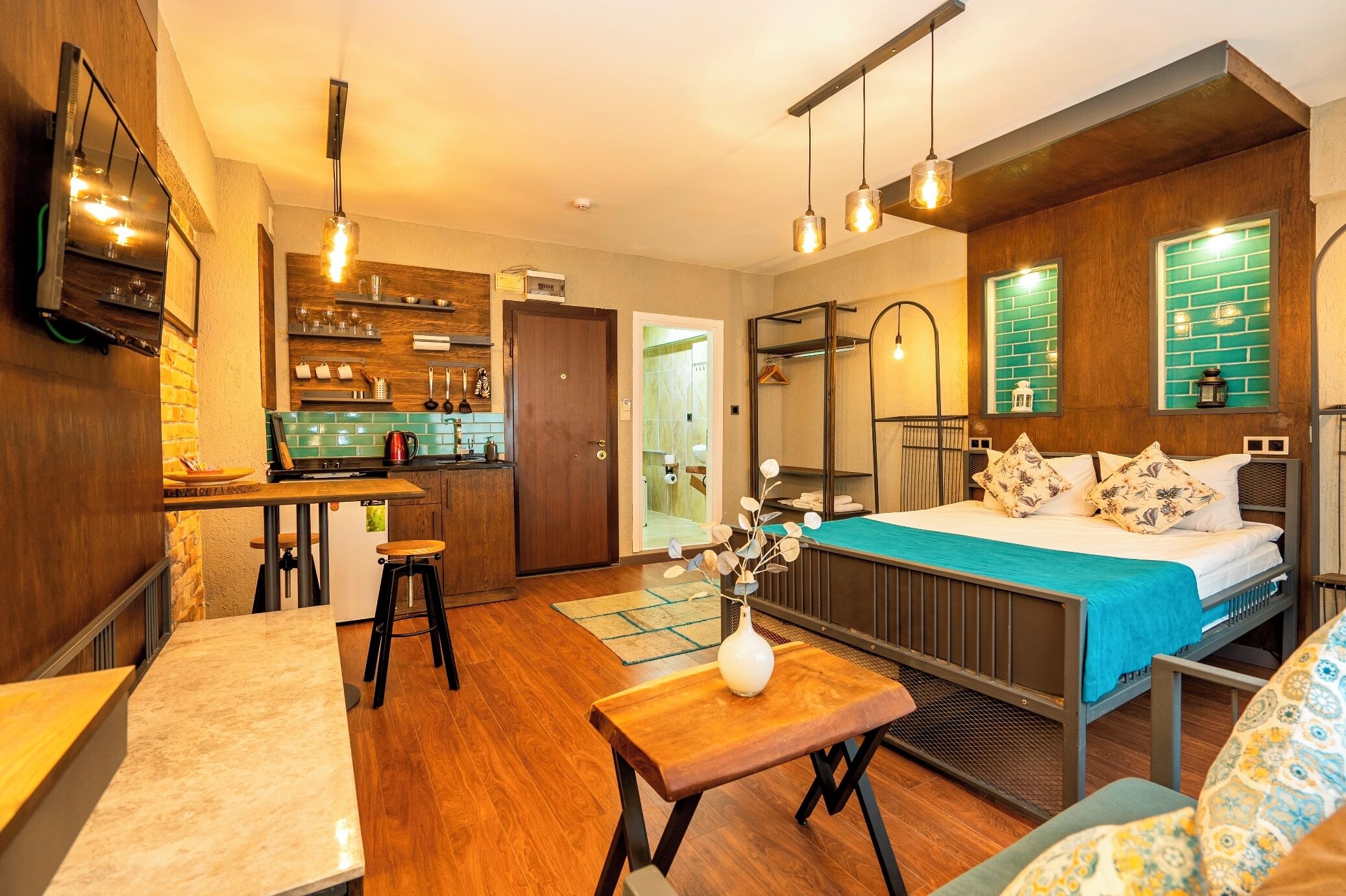 A modern hotel room with a kitchenette, a bed with a blue bedspread, a hanging light fixture, and a wooden floor.