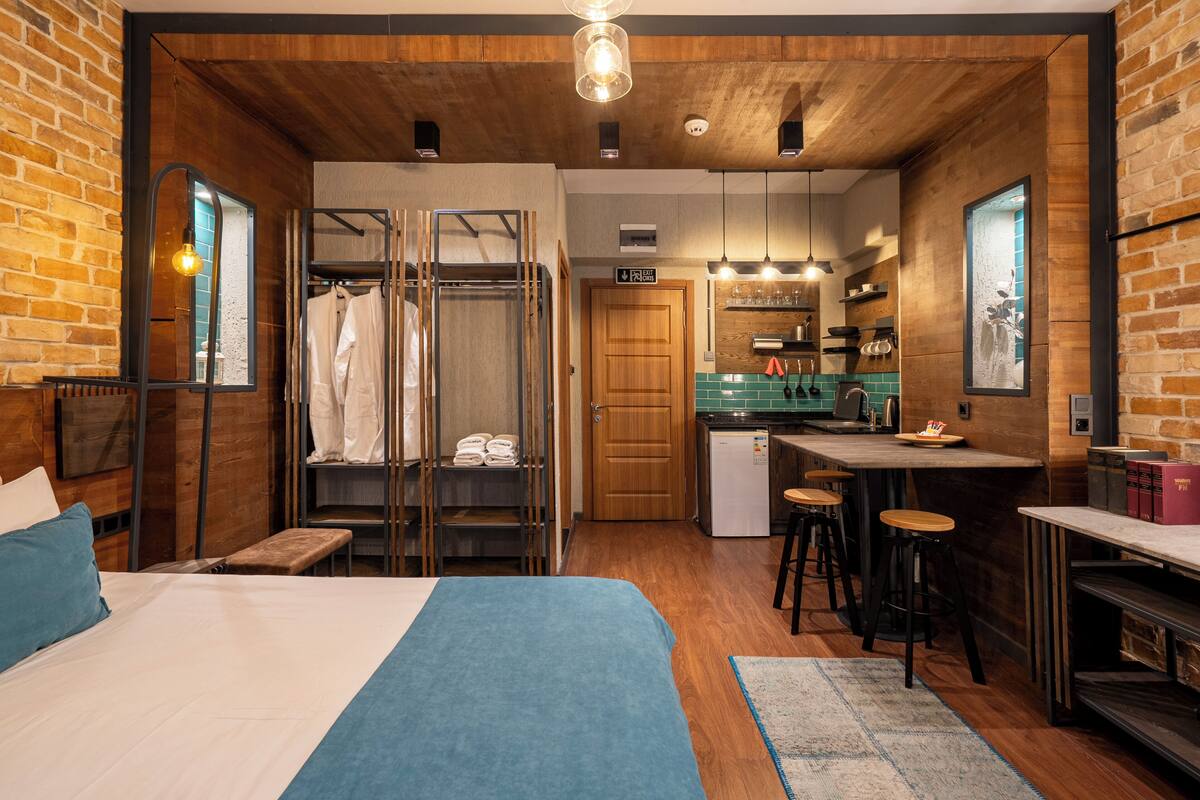 A compact hotel room with a bed, a small kitchenette, a wooden ceiling, and a brick wall.