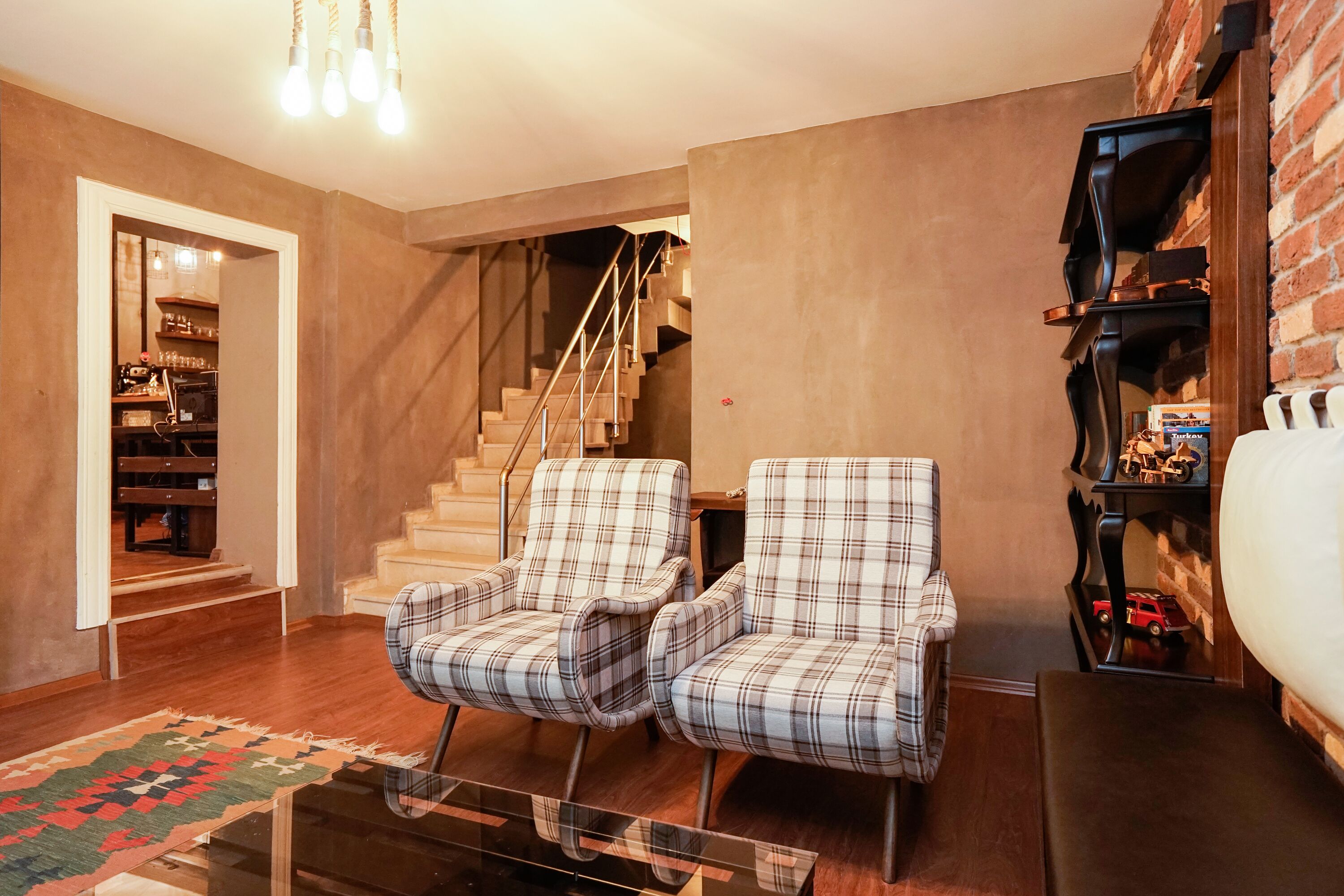 A living room with a staircase, two plaid armchairs, a coffee table, and a brick wall.