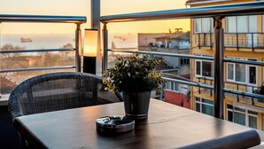 Un balcón con vistas al mar y edificios, una planta en maceta y una mesa con una bandeja.