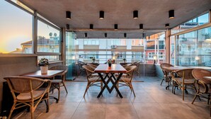 Un área de comedor en la azotea con sillas de mimbre y una mesa central, amplias ventanas con vistas a edificios e iluminación colgante.