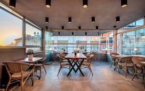 A rooftop dining area with wicker chairs and a central table, large windows offering views of buildings, and pendant lighting.