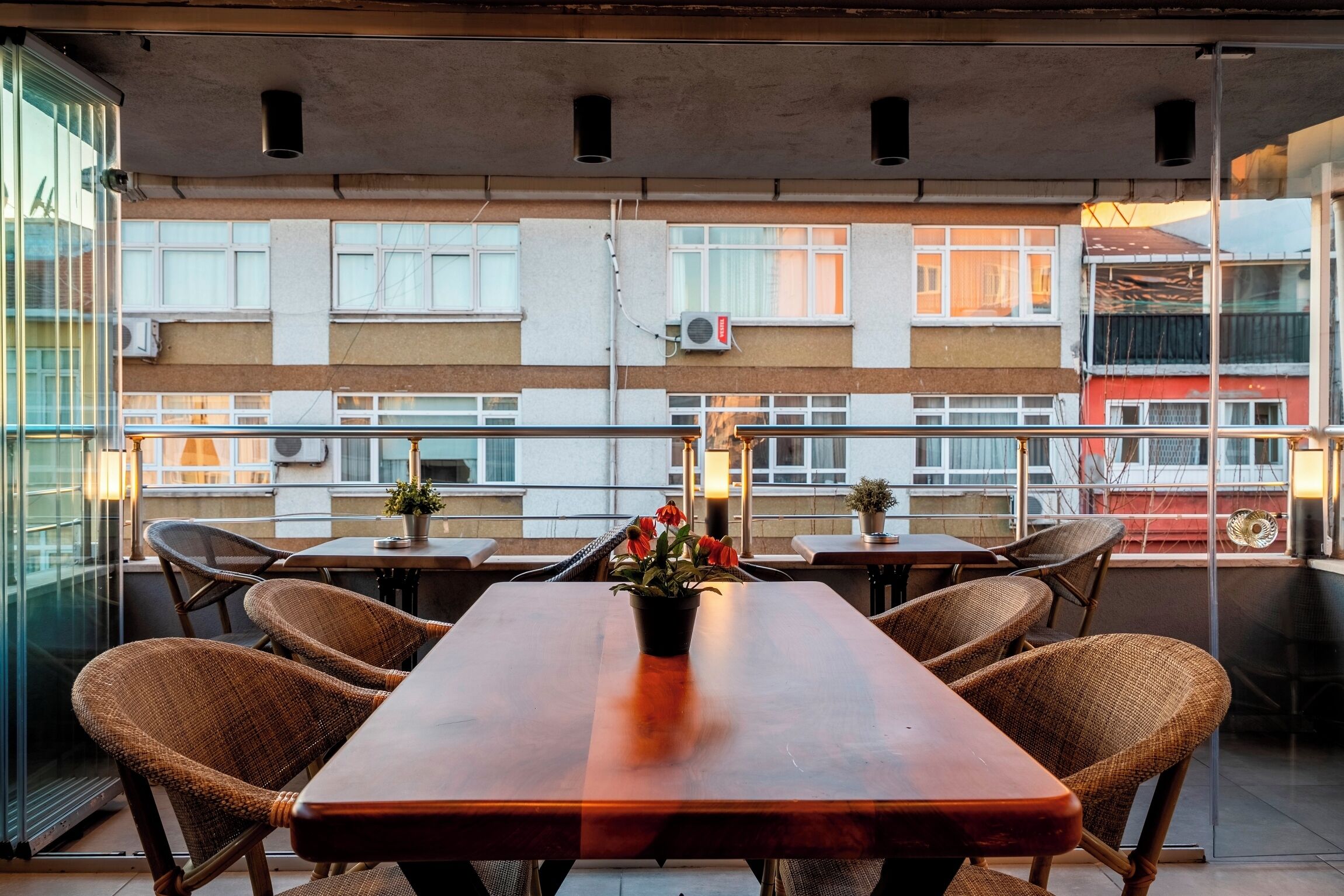 A balcony with a wooden table and wicker chairs, a potted plant, and a view of a building with multiple windows and air conditioning units.