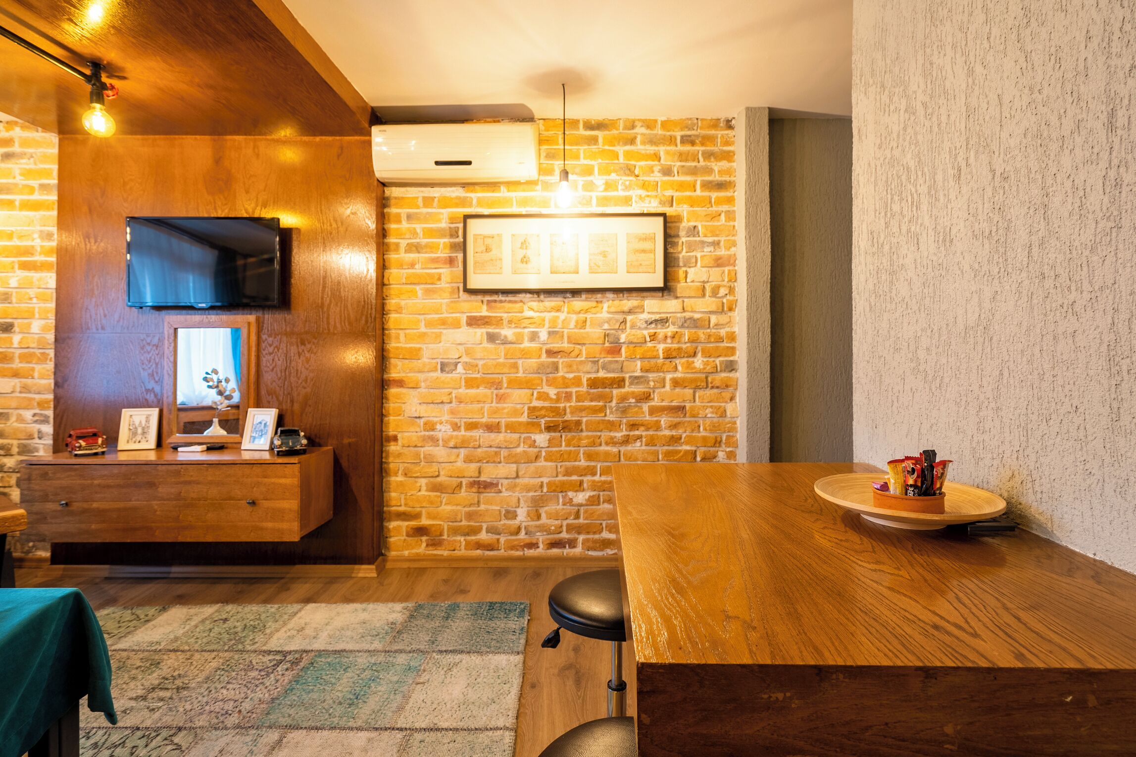 Una habitación con pared de ladrillo, una mesa de madera, un televisor montado en la pared y un cuadro enmarcado.