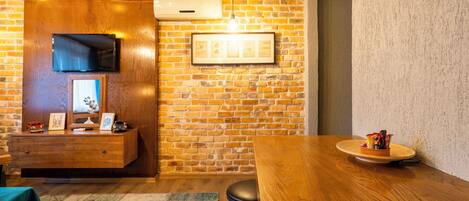 Una habitación con pared de ladrillo, una mesa de madera, un televisor montado en la pared y un cuadro enmarcado.