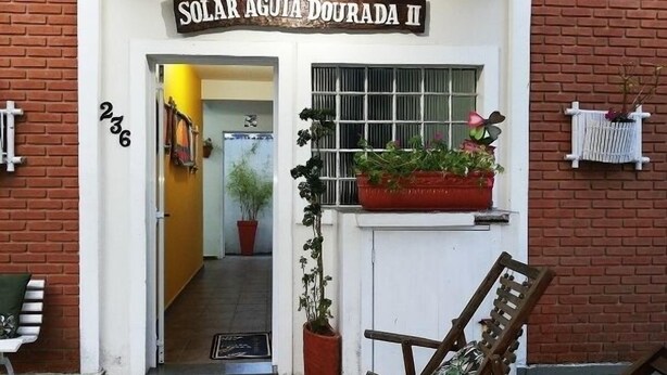 A building entrance with a sign reading "SOLAR  ÁGUA DOUADA II" and a number "236" on the wall. There is a wooden chair outside and a potted plant on the windowsill. - Flat H Ubatuba (Ubatuba)