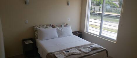 A hotel room with a bed, a nightstand, a window with a view of the street, and a wall-mounted light fixture.