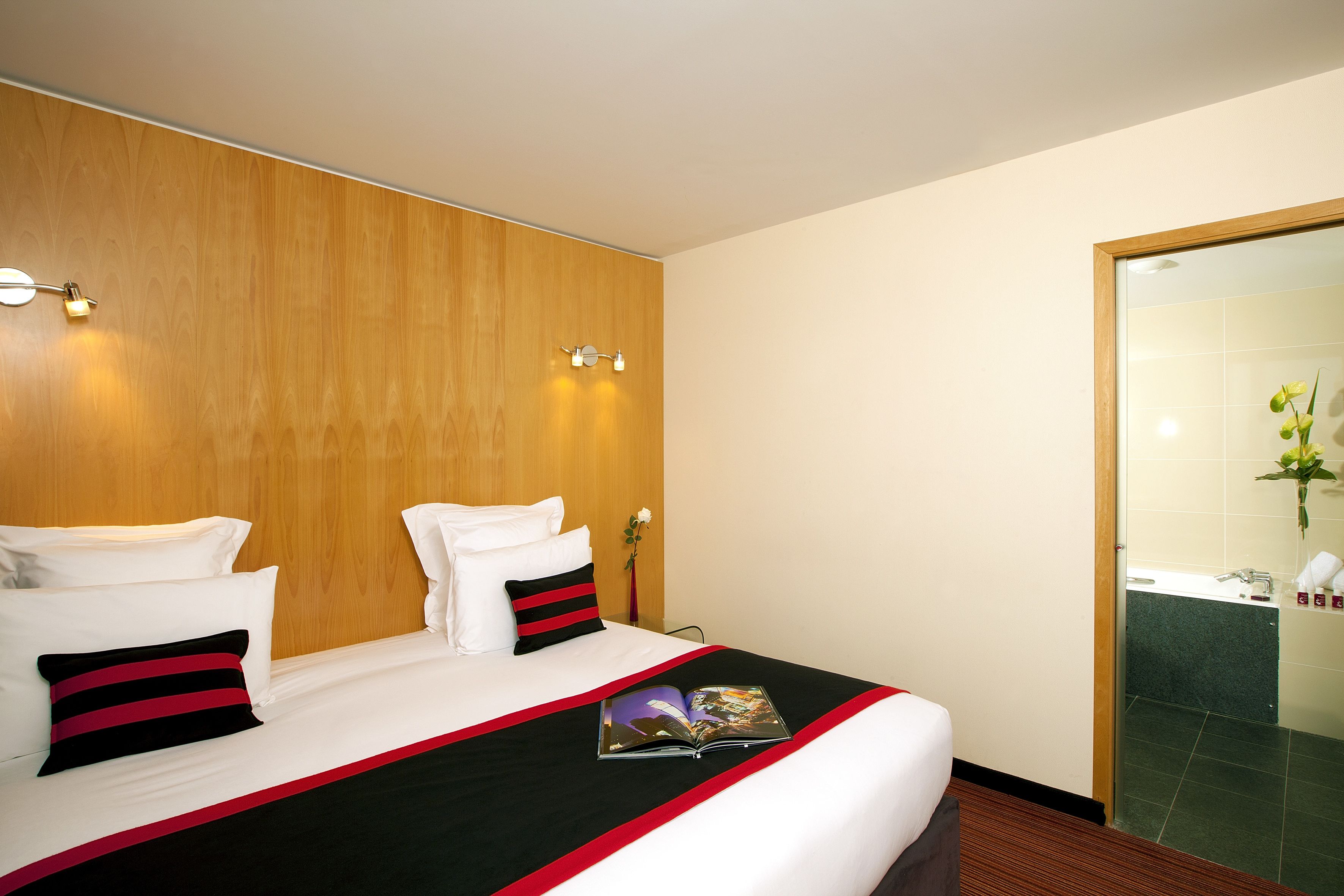 A hotel room with a wooden headboard, two white pillows, a black and red striped pillow, a magazine on the bed, and a bathroom visible through a glass door.
