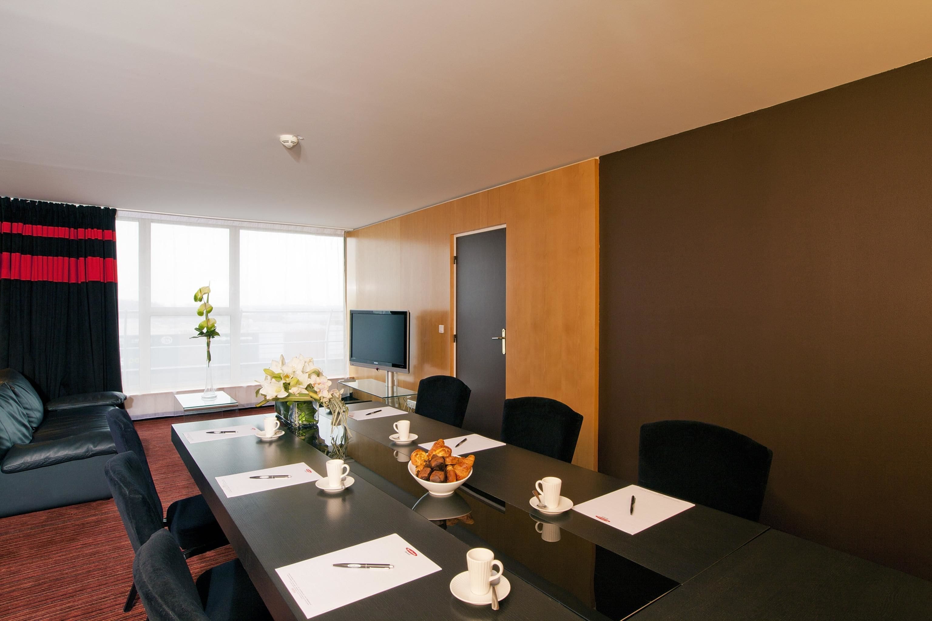 A modern conference room with a long table, black chairs, a TV, and a window with curtains.