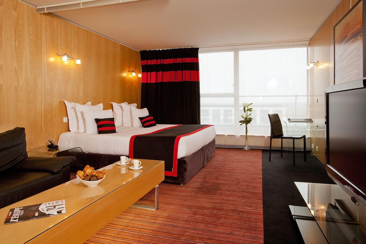 A hotel room with a bed, a TV, a desk with a bowl of food, a coffee cup, a chair, and a window with a view.