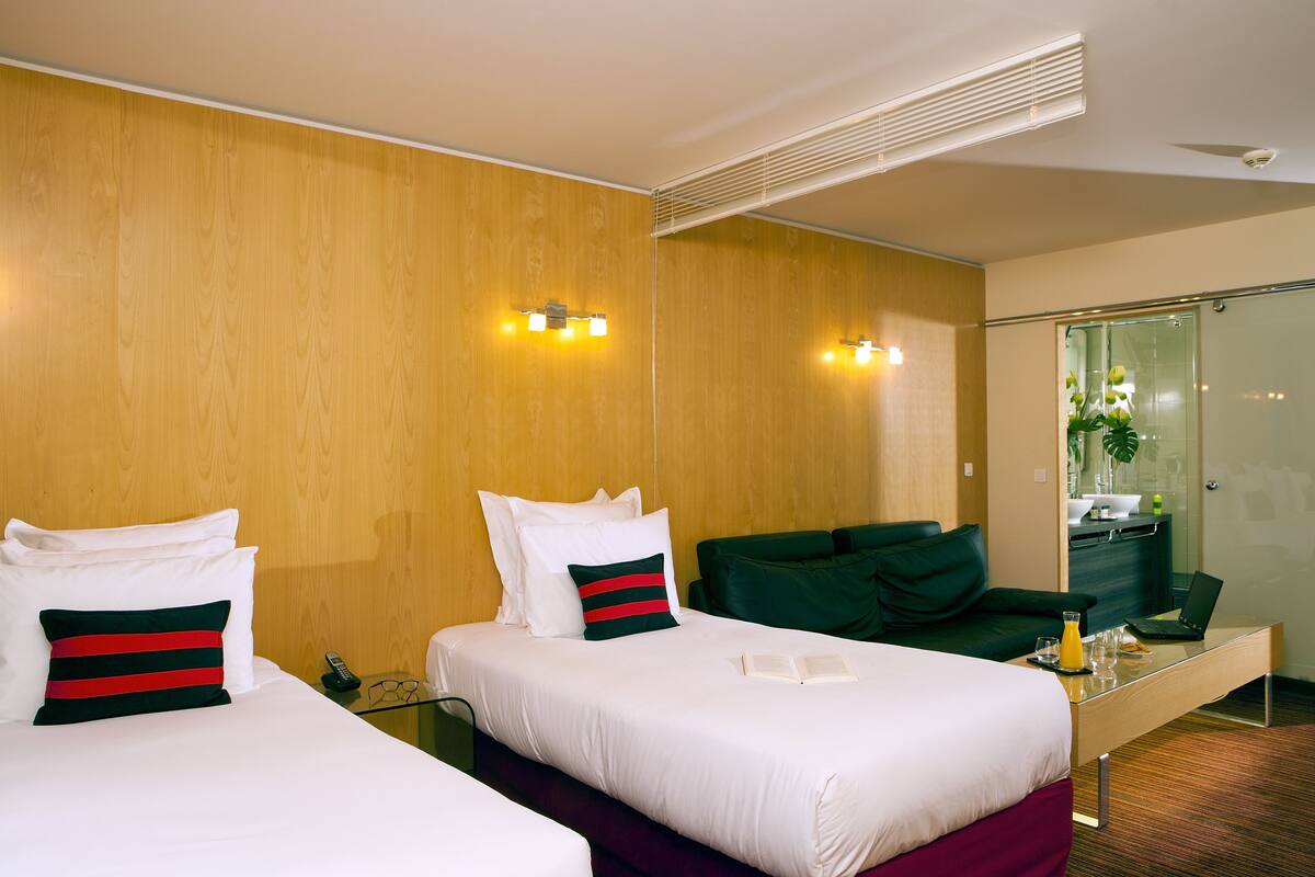 A hotel room with two beds, a desk, a mirror, and a bathroom visible through a glass door.