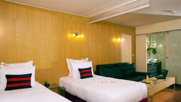 A hotel room with two beds, a desk, a mirror, and a bathroom visible through a glass door.