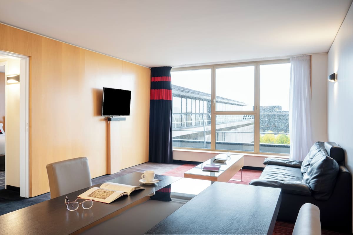 A hotel room with a bed, a desk with a book and glasses, a coffee cup, a TV, and a sofa.