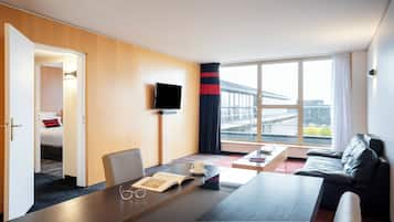 Habitació d'hotel amb un llit, una taula de nit amb un llibre i ulleres, una tassa de cafè, una televisió i un sofà.