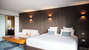 A modern hotel room with a large bed, a sofa, a glass coffee table, and a wooden accent wall with wall-mounted lighting. - Residhome Suites Paris Sénart (Lieusaint)