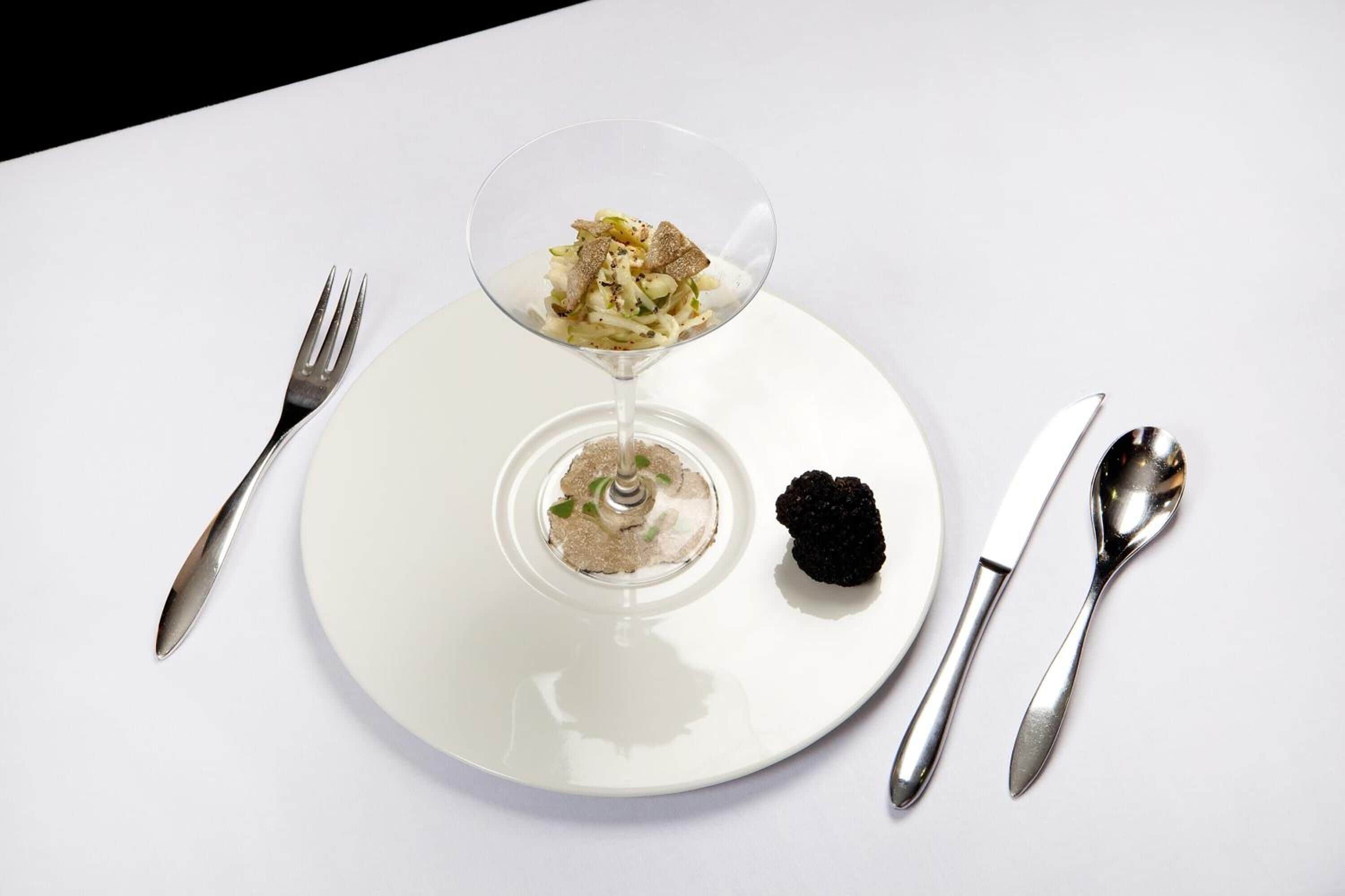 En vit tallrik med en dessert i ett glas, tillsammans med en gaffel, kniv och sked.