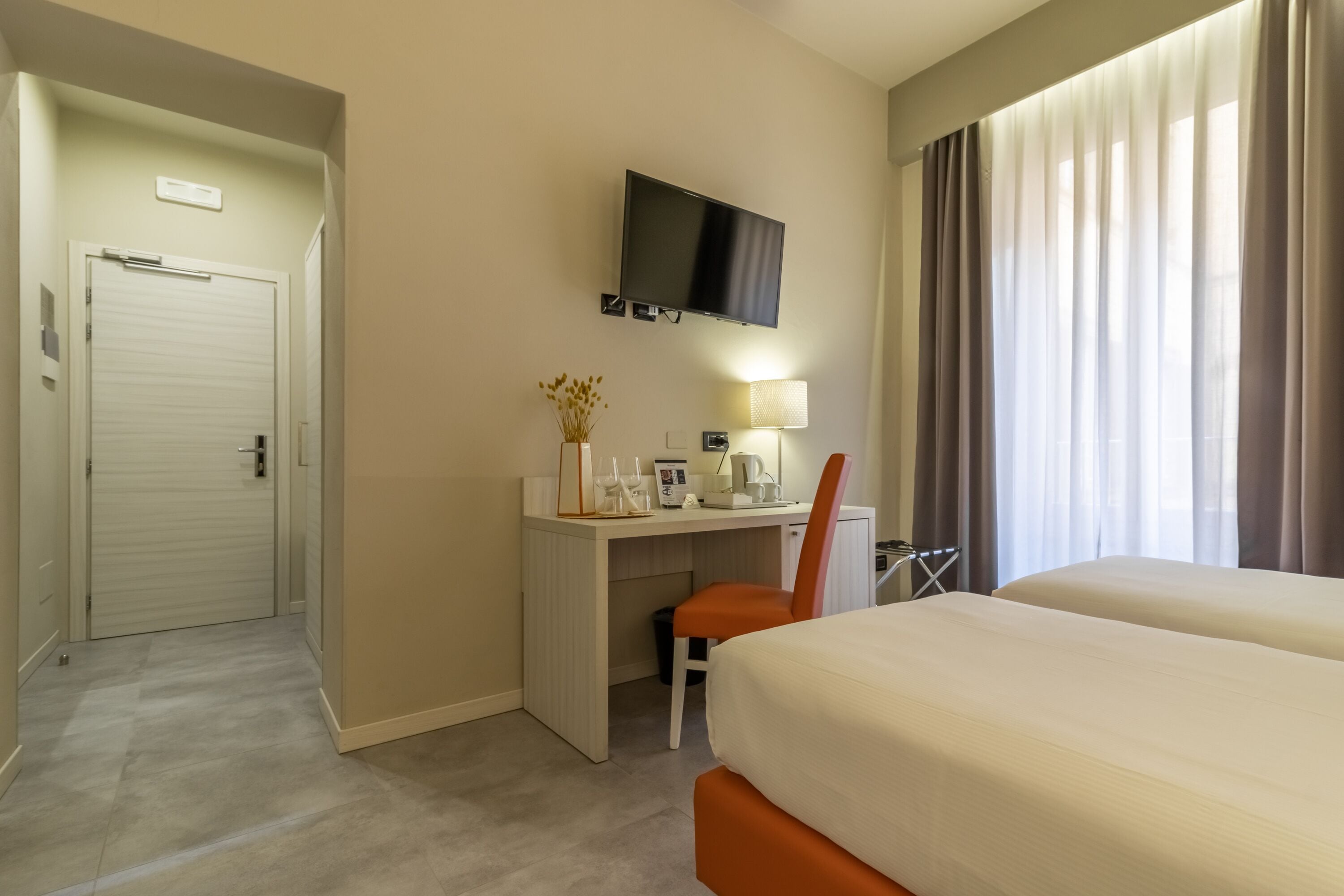 A hotel room with a bed, a desk with a chair, a TV mounted on the wall, and a window with curtains.