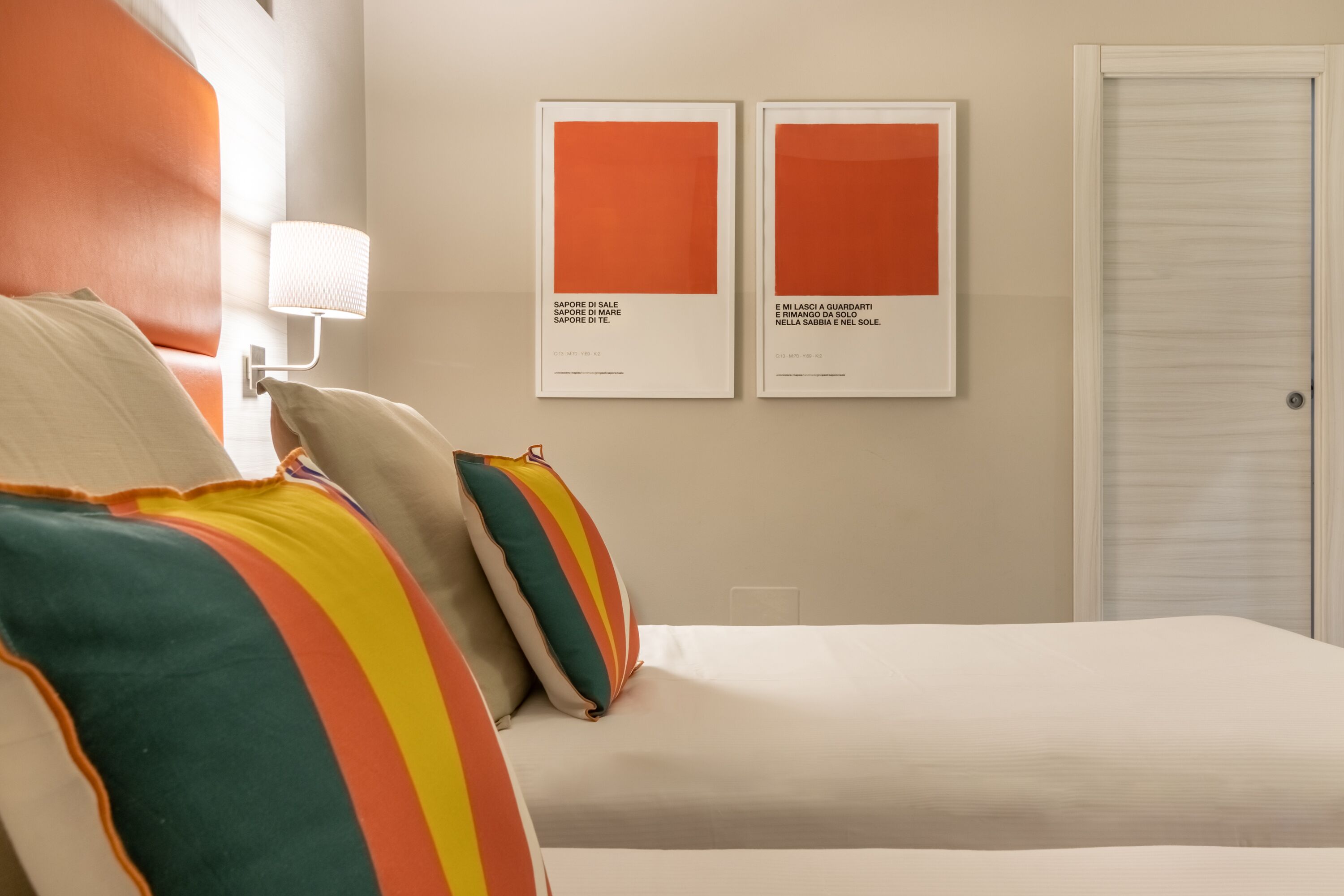 A hotel room with a bed, pillows, a wall-mounted lamp, and two framed artworks.